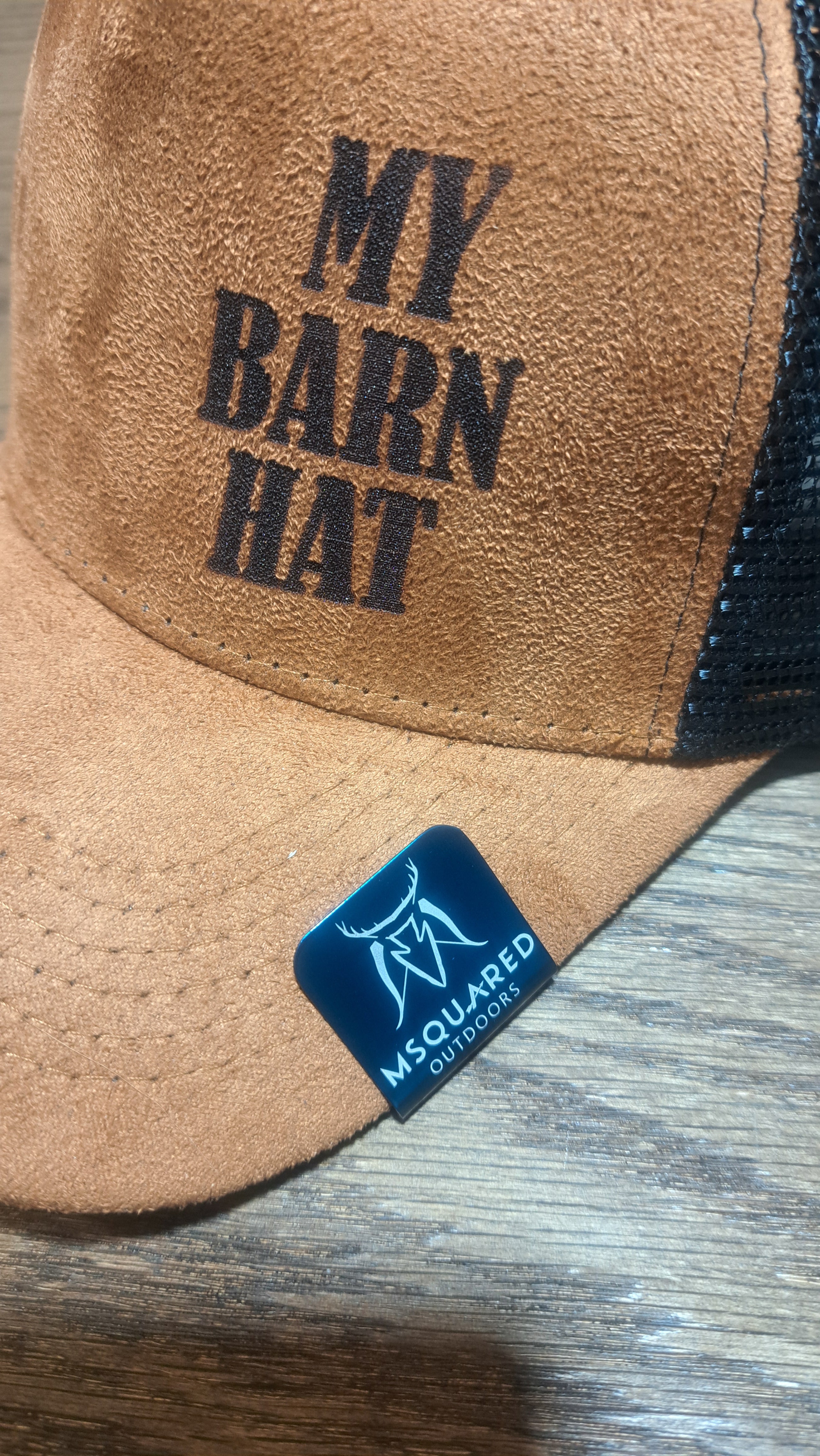 Close-up of a brown leather cap with 'My Barn Hat' text and a blue hat clip with MSquared outdoors engraved on it. 